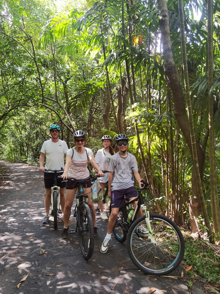 Family cycling on a shaded pathway surrounded by towering trees and lush jungle vegetation