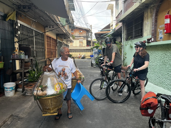 Friendly local vendor carrying his goods on a traditional shoulder pole greeting cyclists in a narrow Bangkok alley