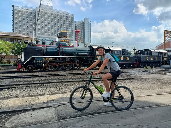 Cyclist posing with bike in front of a historic black steam locomotive at the old Bangkok railway station