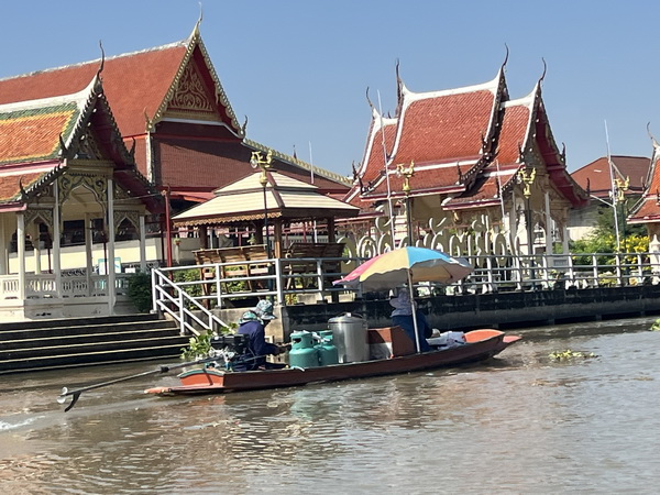 Vendor boat with gas canisters passing a traditional Thai temple by the water