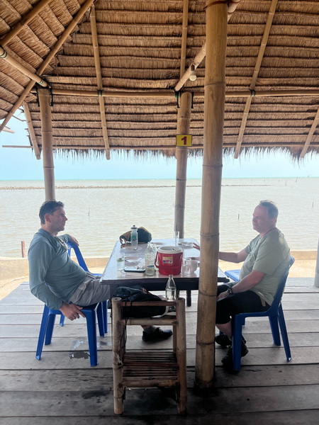 Cyclists enjoying lunch at a tranquil thatched-roof restaurant right on the water's edge