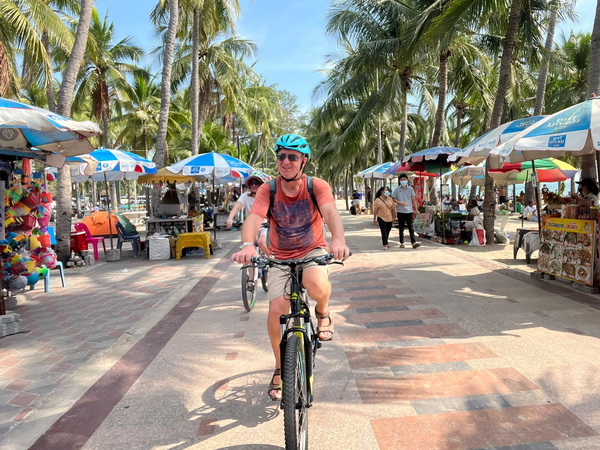Cyclist riding along Bang Saen beach promenade lined with palm trees and colorful market stalls