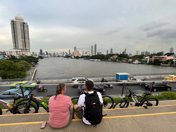 Two cyclists sitting on steps admiring the panoramic Chao Phraya river and city skyline view from the Sky Bridge