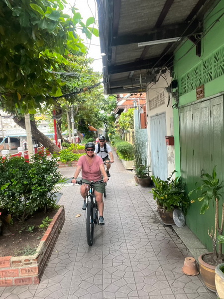 Cyclists riding through a charming narrow canal-side alleyway lined with greenery and traditional houses