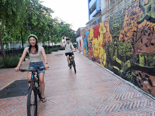 Cyclists riding past a vibrant wall of colourful street art and graffiti in a quiet Bangkok pathway